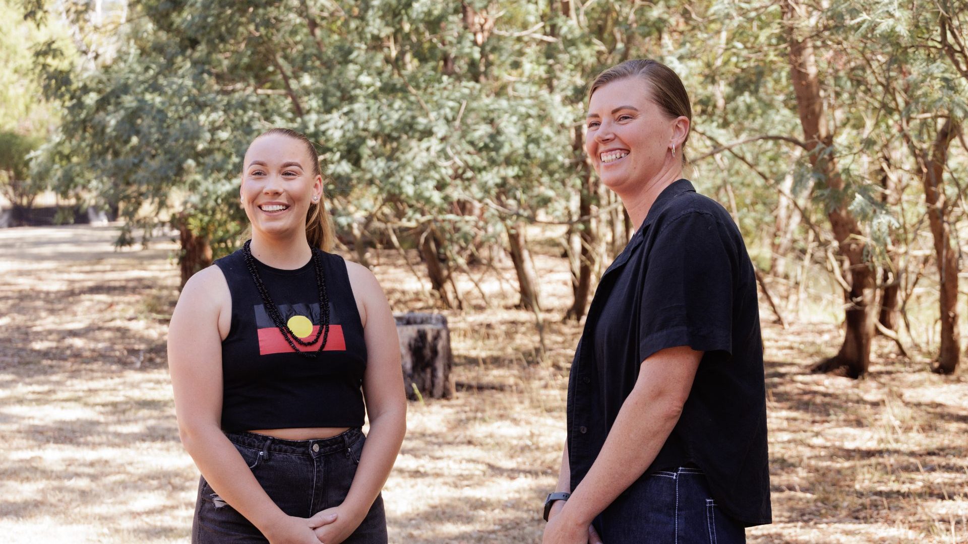 Two women, one with a dark-haired ponytail, other with short camera, smile and look sideways to camera.