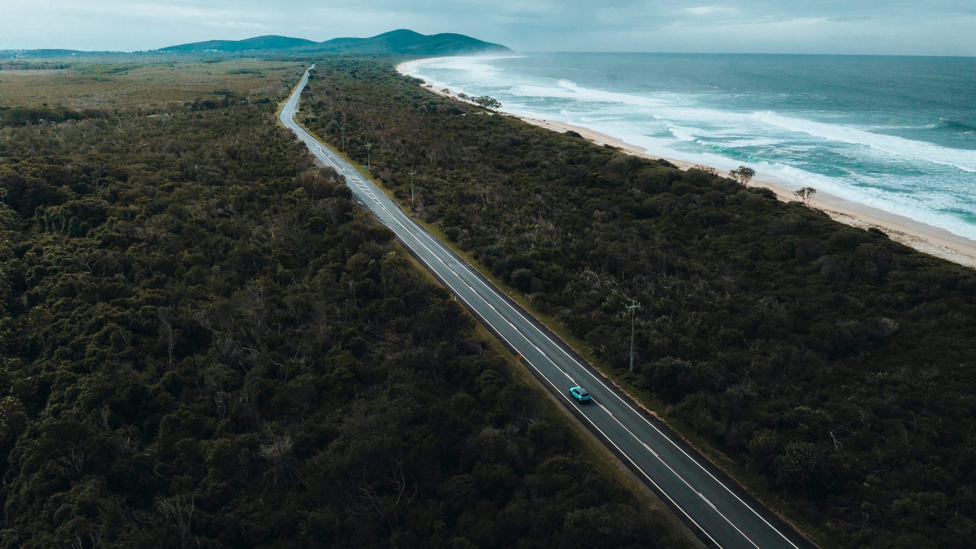 Aerial shot of car driving stretch of road, forest to left and water to right