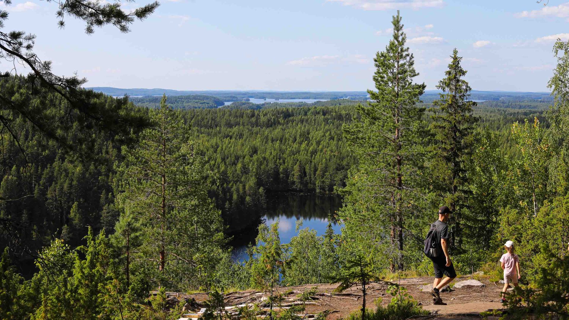 A man and child hike along a forest trail that looks down over a lake.