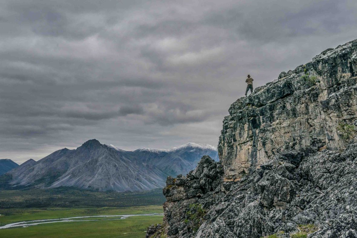 A person standing on the edge of a cliff looks out over mountains and valleys.