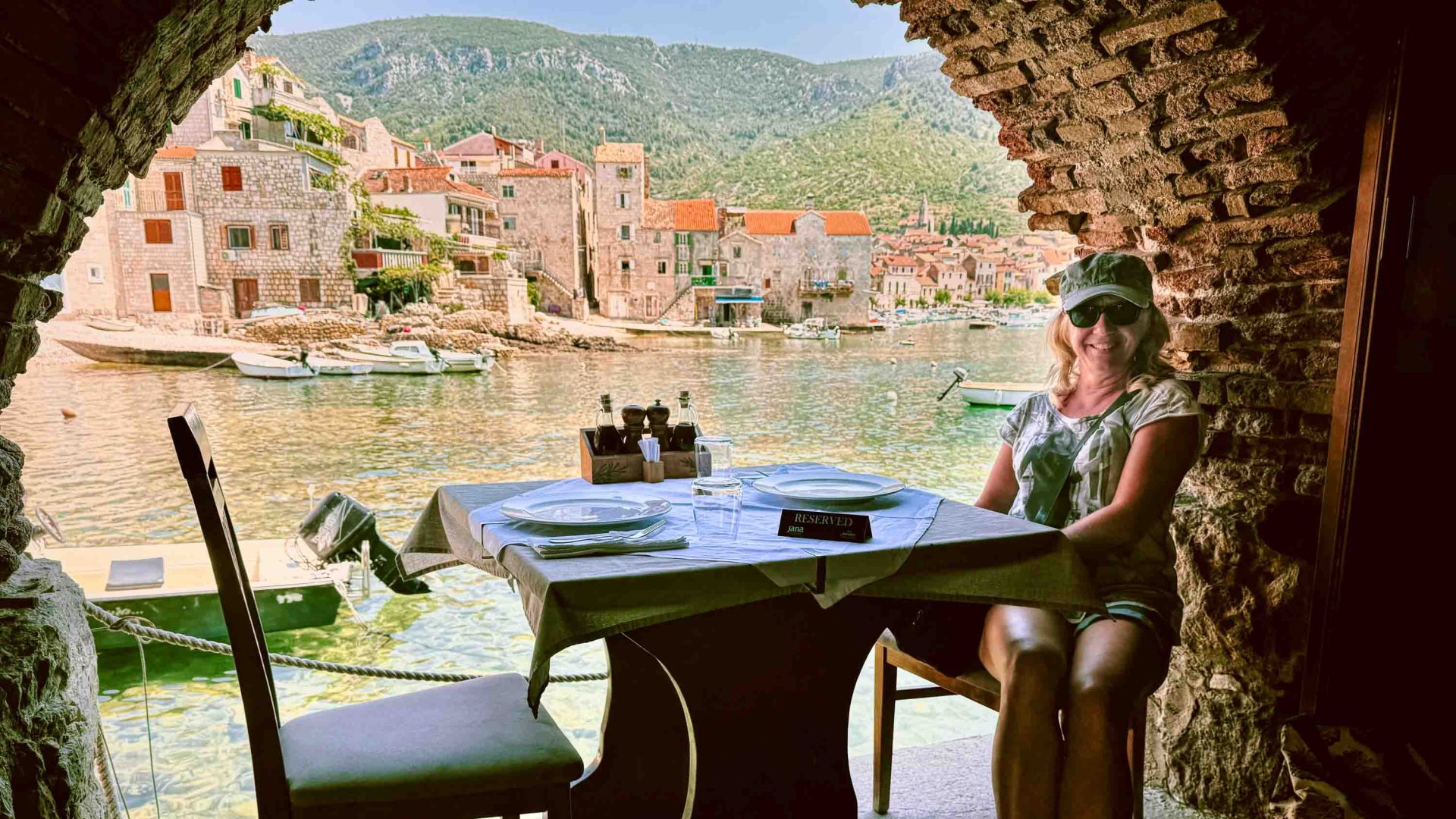 A woman sits at a restaurant table below an arch, with views of a town.