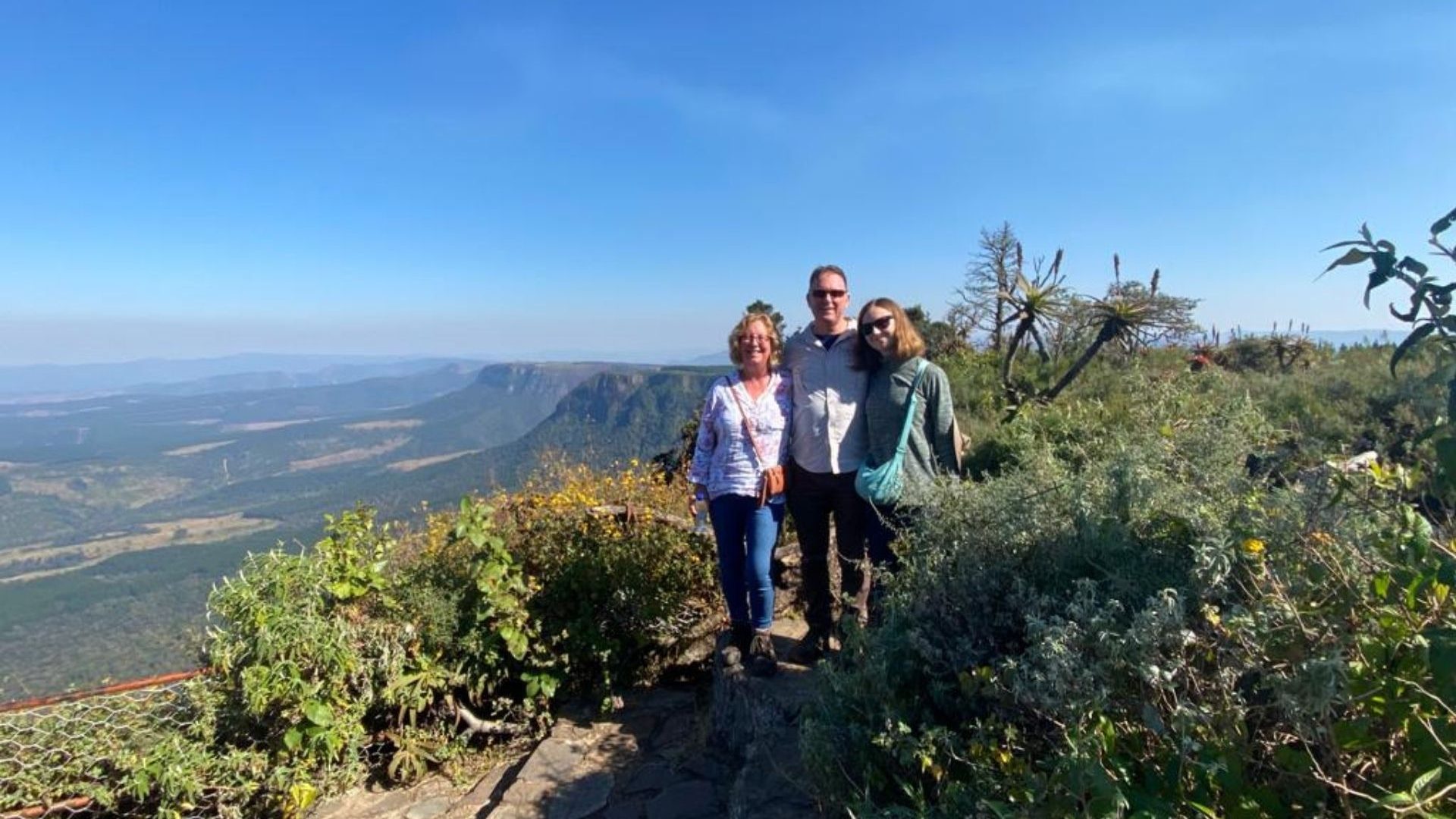 A young girl and two parents pose for a photo atop a viewpoint with mountains and rolling plains behind them.