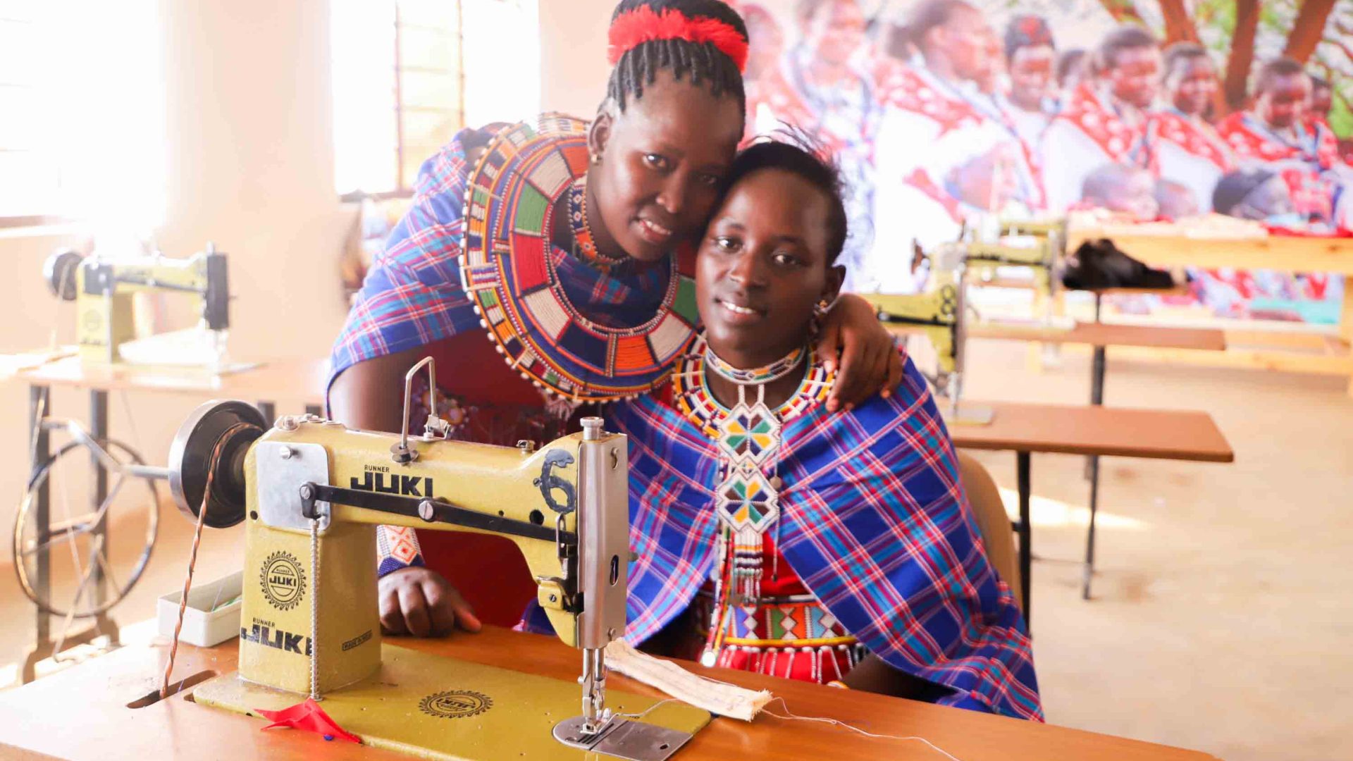 Two women smile to camera while sitting behind a sewing machine.