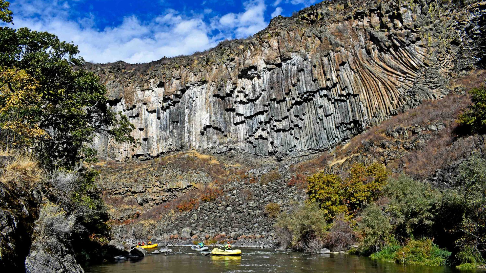 A yellow white water raft goes down a river with tall rocky walls and plants on the edges.