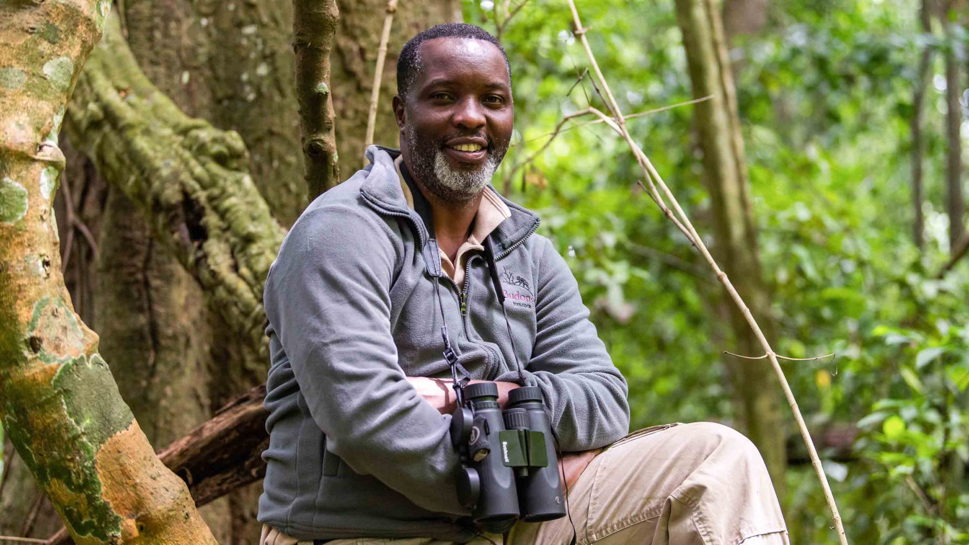 A man sits in front of trees with binoculars round his neck.