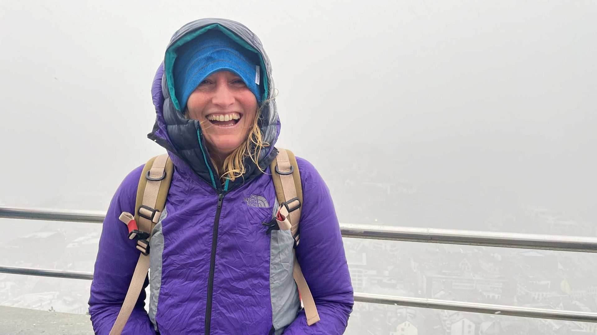 A woman in a purple parker smiles against a foggy mountain background.