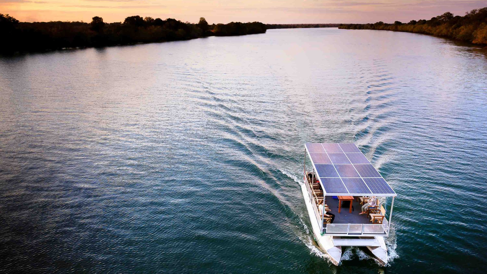 A boat with solar panels on its roof glides through a lake at sunset.
