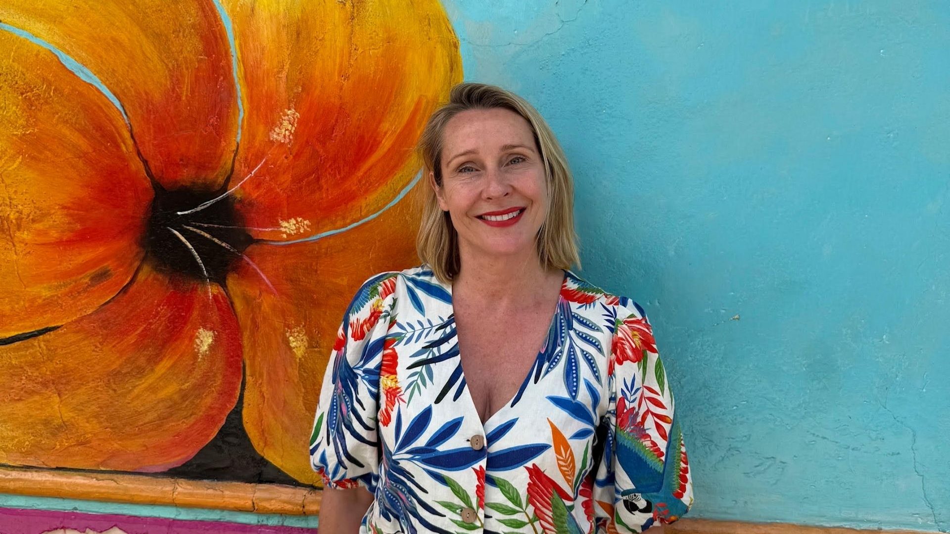 A woman in a colorful printed dress smiles, standing against a blue wall with a yellow flower motif
