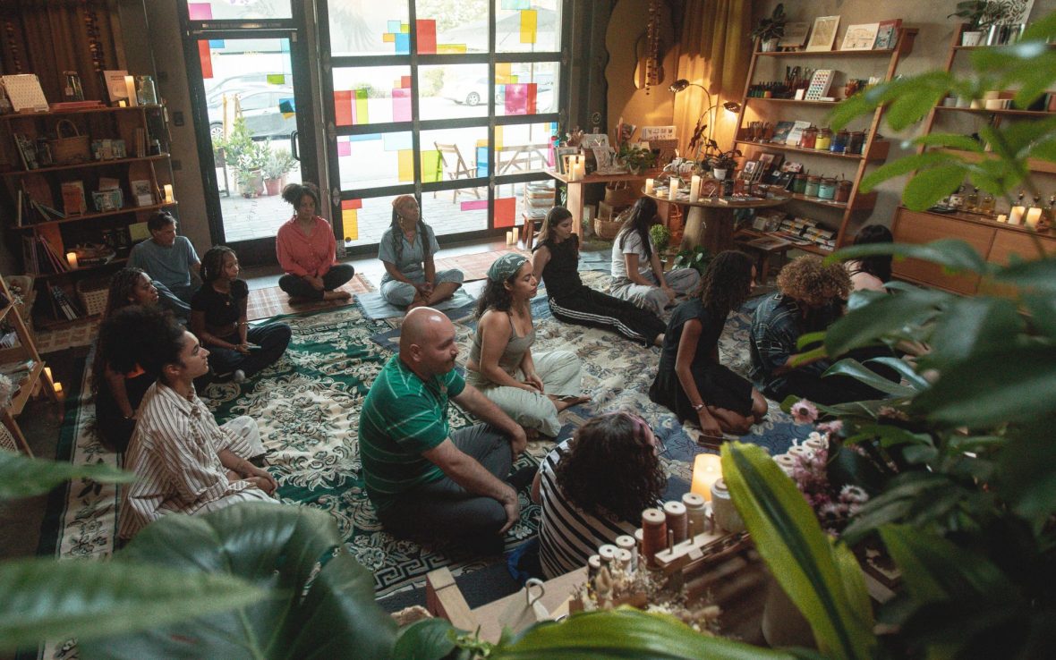 Group of people gather in cozy store, seated on blue carpet, listening to meditation speaker.