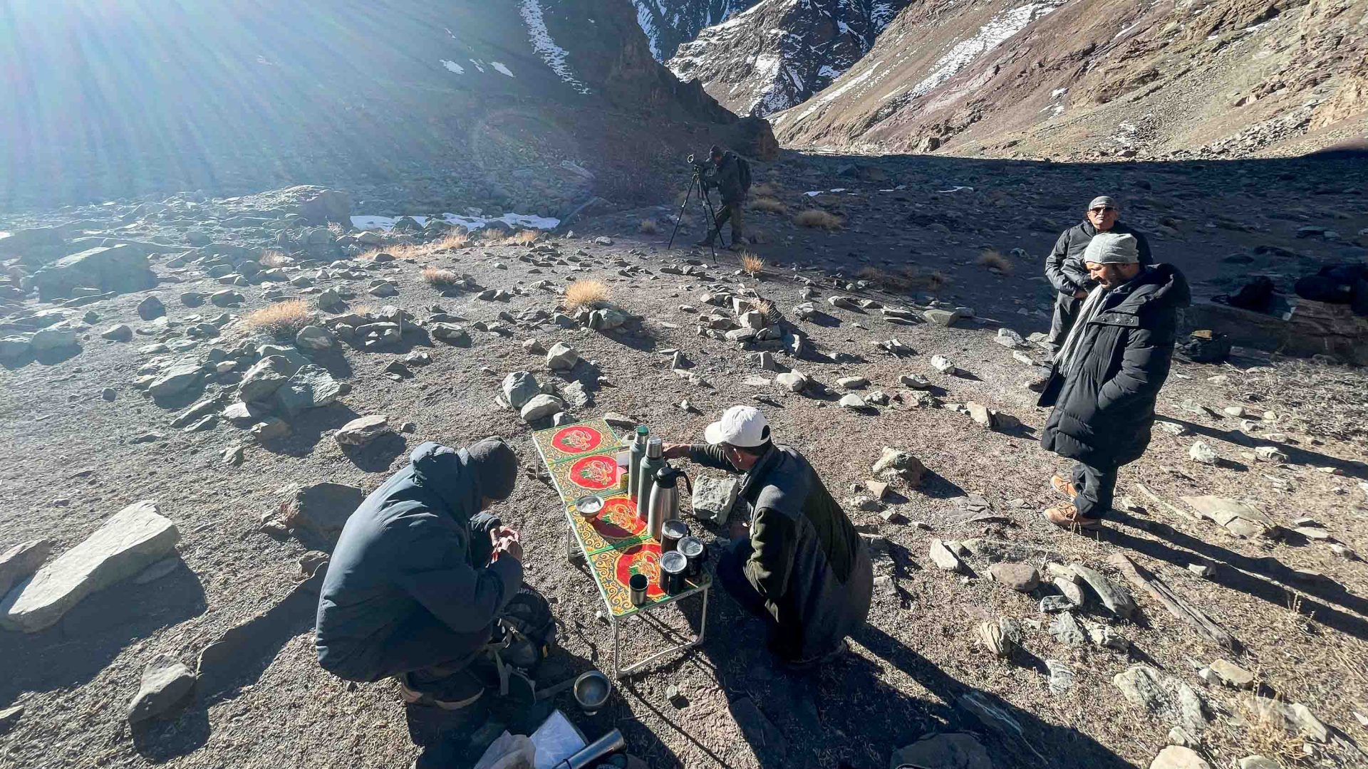 A group rests with tea at a small table. One single person looks out at the mountains with a scope.