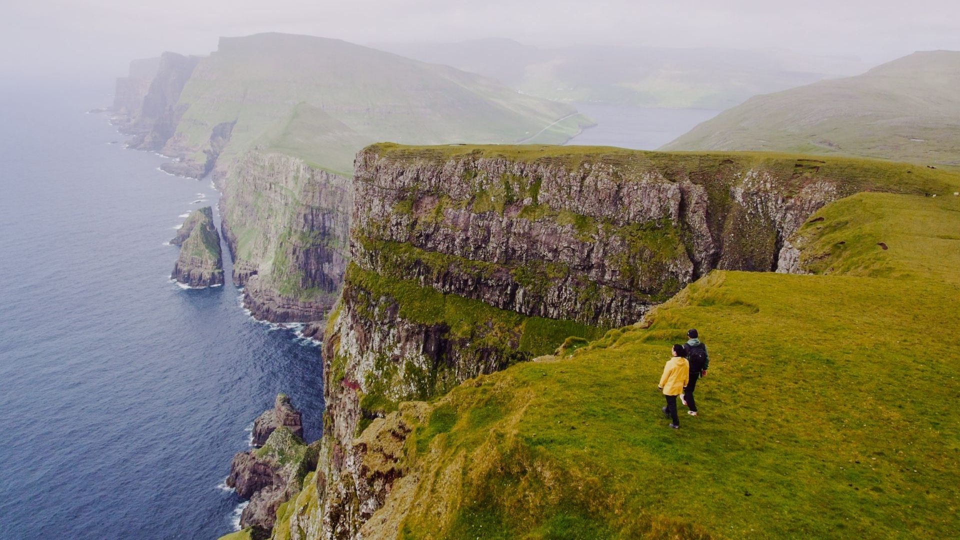 A hiker in a yellow jacket stands near the edge of a grassy cliff overlooking the ocean, with mountains visible in the distance.